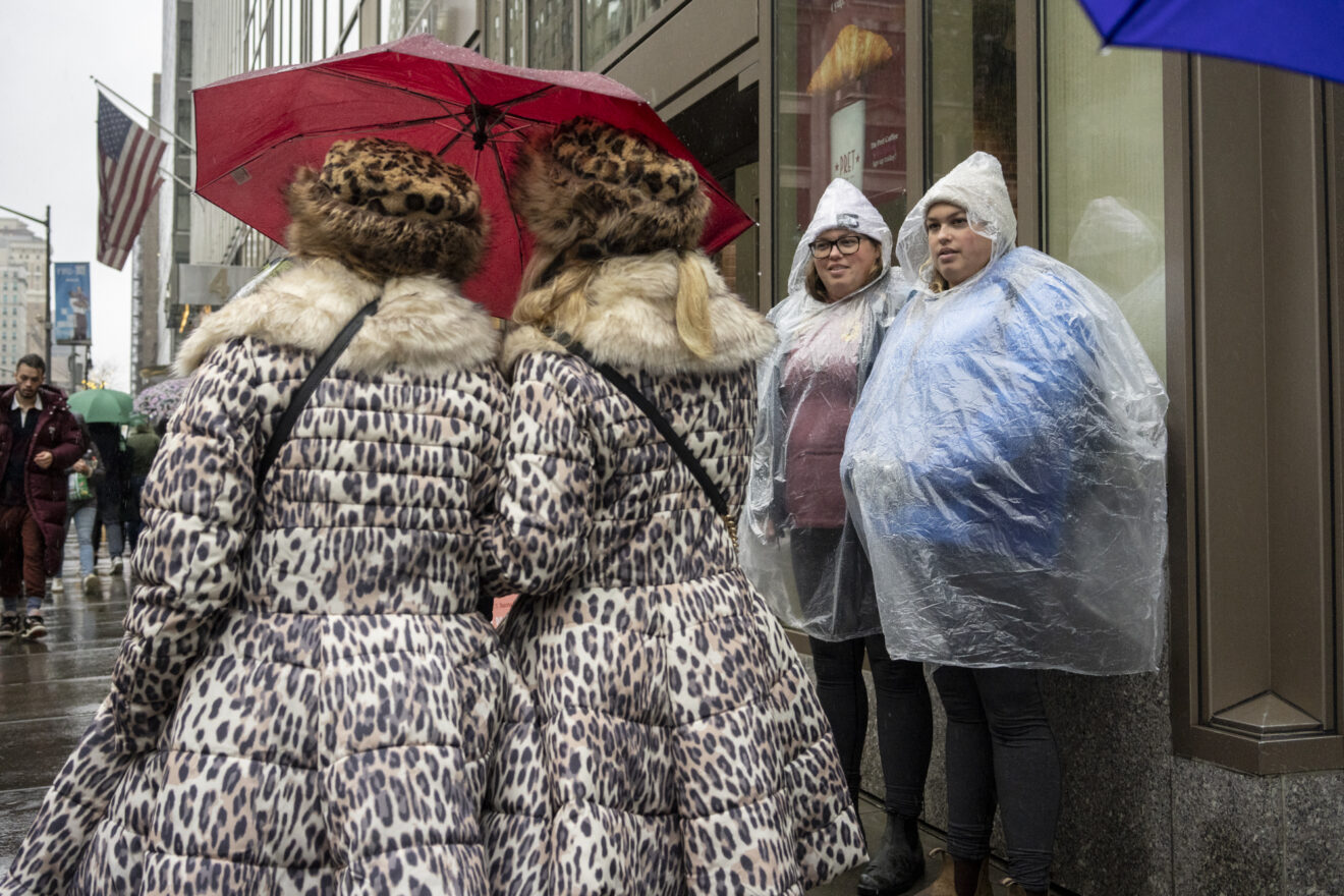Two women seen from behind in matching leopard-print puffer coats and leopard fur hats under a single red umbrella, facing two more women in transparent plastic rain ponchos against a wet Fifth Avenue storefront
